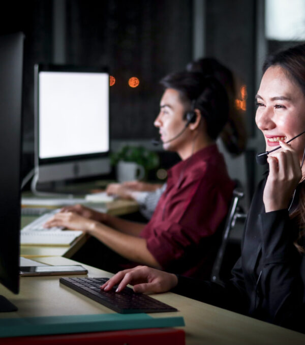 Happy smiling young beautiful Asian woman with headphones working at call center service desk consultant with her teammates at night, ready to take with customer on hands-free phone, happy workplace concept
